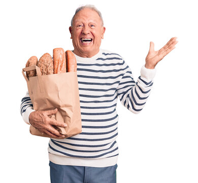 Senior handsome grey-haired man holding paper bag with bread celebrating victory with happy smile and winner expression with raised hands