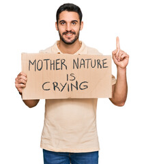 Young hispanic man holding mother nature is crying protest cardboard banner surprised with an idea or question pointing finger with happy face, number one