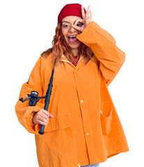 Young latin woman wearing fisher raicoat holding rod smiling happy doing ok sign with hand on eye looking through fingers