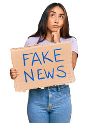 Young latin woman holding fake news banner serious face thinking about question with hand on chin,...