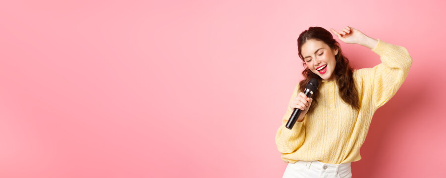 Carefree modern woman dancing and singing song in microphone, performing with mic, smiling and moving relaxed, standing against pink background
