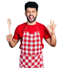 Young arab man with beard wearing baker uniform holding wooden spoon sticking tongue out happy with...