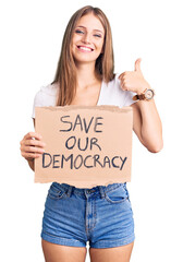 Young beautiful blonde woman holding save our democracy protest banner smiling happy and positive, thumb up doing excellent and approval sign