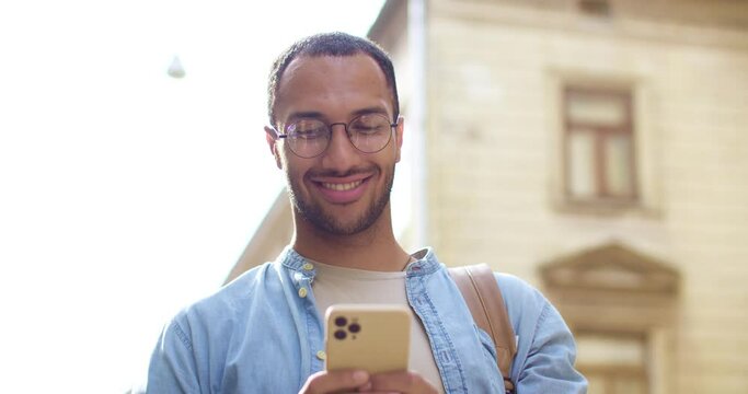 Close up of happy young handsome smiling male torist with backpack texting sms on mobile phone and reading message. Outdoors. Attractive stylish man tapping and scrolling on smartphone.