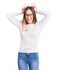 Fototapeta premium Beautiful brunette young woman wearing casual white sweater and glasses posing funny and crazy with fingers on head as bunny ears, smiling cheerful