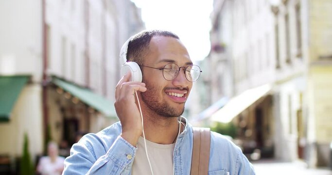 Close up of happy young handsome smiling male torist with backpack and in headphones listening to music and enjoying a day. Outside. Attractive stylish man having musical time and walk.