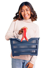 Young beautiful mixed race woman holding supermarket shopping basket looking positive and happy standing and smiling with a confident smile showing teeth