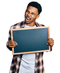 Young african american man holding blackboard angry and mad screaming frustrated and furious,...