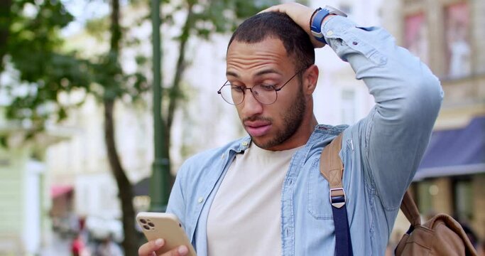 Concerned young handsome man in eyeglasses standing at city street and texting message on smartphone. Outdoor. Good-looking worried and troubled male in town chatting and tapping on mobile phone.