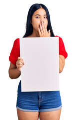 Young beautiful asian girl holding blank empty banner covering mouth with hand, shocked and afraid for mistake. surprised expression