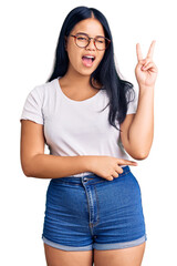 Young beautiful asian girl wearing casual clothes and glasses smiling with happy face winking at the camera doing victory sign with fingers. number two.