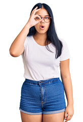 Young beautiful asian girl wearing casual clothes and glasses doing ok gesture shocked with surprised face, eye looking through fingers. unbelieving expression.