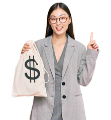 Young chinese woman wearing business suit holding dollars bag surprised with an idea or question pointing finger with happy face, number one