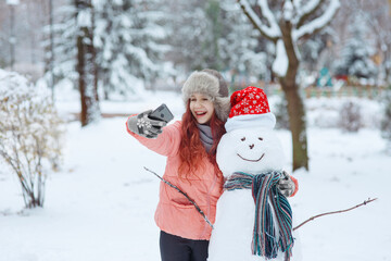Young happy cheerful laughing girl in fur hat hugging a smiling snowman in christmas hat and scarf,...