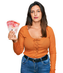 Beautiful hispanic woman holding 20 israel shekels banknotes thinking attitude and sober expression...