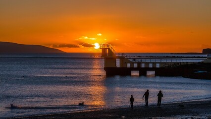 Family walks on sandy beach near Galway Bay at Blackrock Diving Tower during bright orange sunset