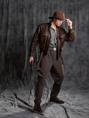 A young man in retro style, an adventure character. The hero of the adventure, a guy in a hat and a leather jacket, posing in the studio on a gray background