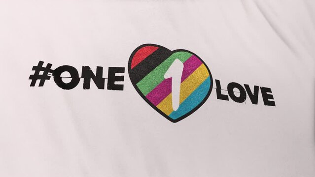 Hashtag symbol "One Love" and rainbow colored heart icon on white rippled flag loop background. 3D animation of LGBT transgender gay, queer rights inclusion in world football, soccer championship.