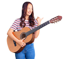 Fototapeta premium Young beautiful chinese girl playing classical guitar pointing thumb up to the side smiling happy with open mouth