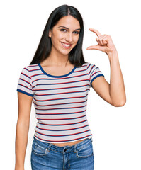 Young hispanic girl wearing casual striped t shirt smiling and confident gesturing with hand doing small size sign with fingers looking and the camera. measure concept.