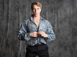 Fototapeta na wymiar Stylish elegant young man in a blue silk shirt posing in the studio on a gray fabric background. sexy pose