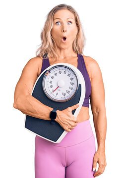 Middle age fit blonde woman wearing sports clothes holding weighing machine scared and amazed with open mouth for surprise, disbelief face