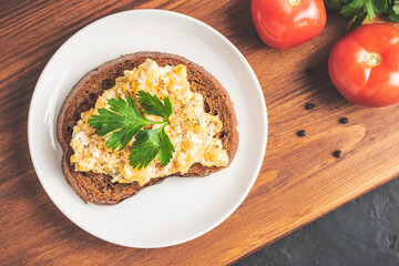 Scrambled eggs with parsley on bread at white plate, homemade healthy breakfast on wooden board, top view