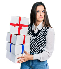 Young brunette teenager holding gifts clueless and confused expression. doubt concept.