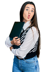 Young brunette teenager hugging laptop with love making fish face with mouth and squinting eyes,...