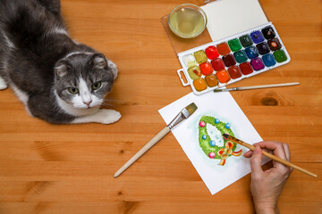 An unrecognizable woman painting Christmas garland at her home with her cat watching her work