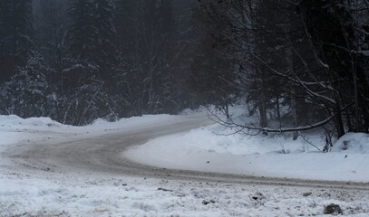 A poorly plowed winter country road, leading through a foggy and tree lined mountain road.