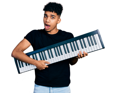Young african american man holding piano keyboard in shock face, looking skeptical and sarcastic, surprised with open mouth