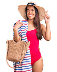 Young beautiful latin girl wearing swimwear and summer hat holding beach towel and bag surprised with an idea or question pointing finger with happy face, number one