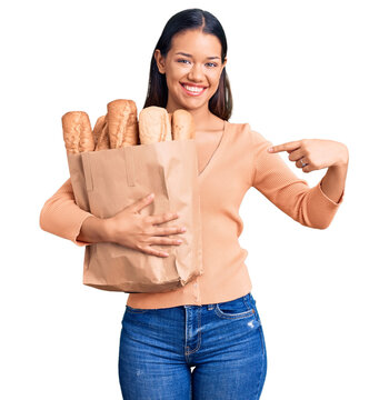Young beautiful latin girl holding paper bag with bread pointing finger to one self smiling happy and proud