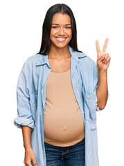 Beautiful hispanic woman expecting a baby showing pregnant belly smiling with happy face winking at...