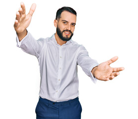 Naklejka premium Young man with beard wearing business shirt looking at the camera smiling with open arms for hug. cheerful expression embracing happiness.
