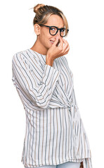 Beautiful blonde woman wearing business shirt and glasses smelling something stinky and disgusting, intolerable smell, holding breath with fingers on nose. bad smell