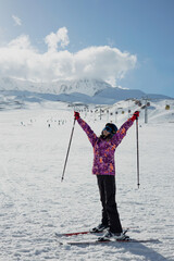 A girl on alpine skis dressed in a ski suit and helmet stands against the backdrop of snow-covered mountains and a bright blue sky.  Winter. Sport and travel content  