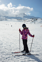 A girl on alpine skis dressed in a ski suit and helmet stands against the backdrop of snow-covered mountains and a bright blue sky.  Winter. Sport and travel content  