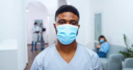 Fototapeta na wymiar Close up portrait of African American young male doctor in medical mask standing in hospital hall and looking at camera. Mixed-races healthcare professionals on background Coronavirus epidemic concept