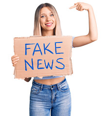 Young beautiful blonde woman holding fake news banner pointing finger to one self smiling happy and...