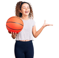 Beautiful kid girl with curly hair holding basketball ball celebrating victory with happy smile and...