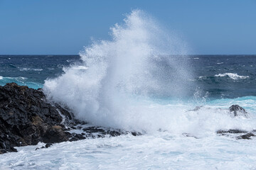 Fototapeta premium a big wave breaks against some rocks on the seashore, on a rough sea day, foam from the waves on the blue ocean, powerful splash of waves breaking against the sharp rocks