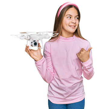 Beautiful brunette little girl using drone pointing thumb up to the side smiling happy with open mouth
