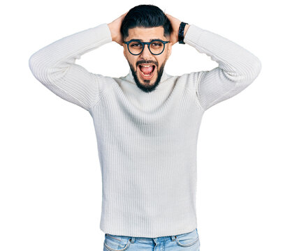 Young arab man with beard wearing elegant turtleneck sweater and glasses crazy and scared with hands on head, afraid and surprised of shock with open mouth