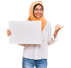 Young brunette arab woman wearing traditional islamic hijab holding banner pointing thumb up to the side smiling happy with open mouth