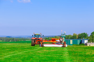 A large rotary mower attached to a tractor mows the grass in the field near the building.