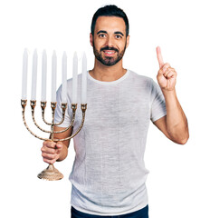 Young hispanic man with beard holding menorah hanukkah jewish candle smiling with an idea or...