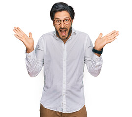 Naklejka premium Young hispanic man wearing business shirt and glasses celebrating crazy and amazed for success with arms raised and open eyes screaming excited. winner concept
