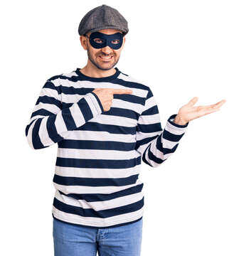 Young handsome man wearing burglar mask amazed and smiling to the camera while presenting with hand and pointing with finger.